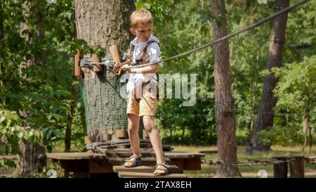 Little boy holding safety rope and walking over wobbly wooden bridge at adventure park. Active childhood, healthy lifestyle, kids playing outdoors, ch Stock Photo
