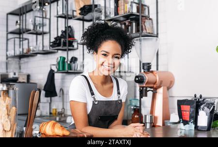 Beautiful female barista looking at camera. Cheerful waitress in an apron standing at a counter in a cafe. Stock Photo