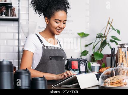 Barista in apron typing on digital tablet. Cheerful woman with curly hair working as a barista in a coffee shop using a digital tablet. Stock Photo