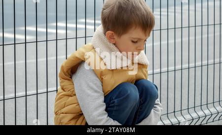 Upset lonely boy being bullied at school sitting next to metal fence at playground. Child depression, problems with bullying, victim in school, emigra Stock Photo