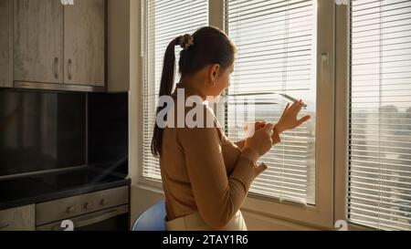 Young brunette woman drinking coffee and looking on city through window blinds. Stock Photo