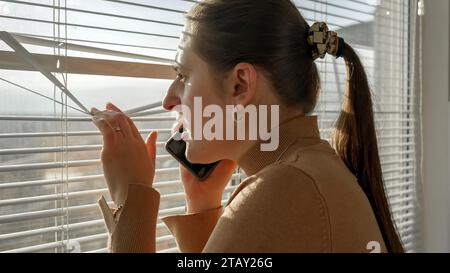 Young scared woman looking through window on street and calling 911 police. Crime witness, spying through window, peeking on street Stock Photo