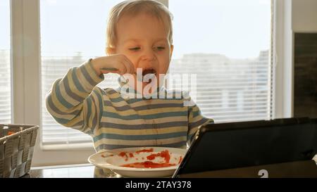 Little boy getting dirty and messy while eating soup on kitchen with tablet computer. Domestic food, gadget addiction, children healthy nutrition Stock Photo