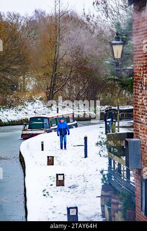 Man walking on canal  towpath past narrowboat moored on the Trent and Mersey canal in the snow during winter at Wheelock  Cheshire England UK Stock Photo