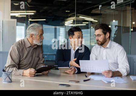 Three diverse business people talking together sitting at a meeting desk inside office, businessmen in business suits discussing with boss business owner about financial indicators, achievements. Stock Photo