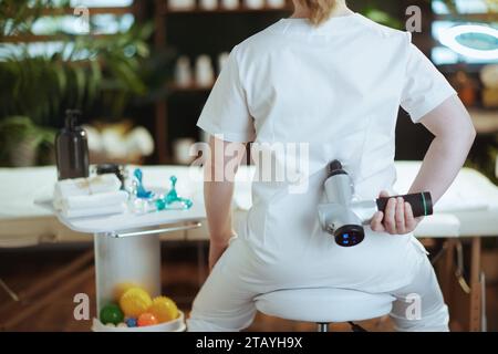Healthcare time. Seen from behind massage therapist woman in spa salon with massage pistol. Stock Photo