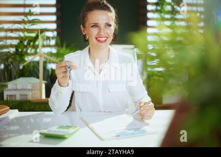 Eco real estate business. happy elegant 40 years old woman realtor in modern green office in white blouse with calculator, clipboard, document and key Stock Photo
