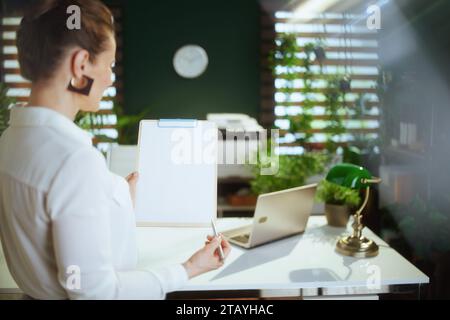 Time to move on. Seen from behind young female real estate agent in modern green office in white blouse with clipboard. Stock Photo