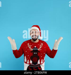 Happy young man in Christmas sweater, Santa hat and knitted mittens on light blue background Stock Photo