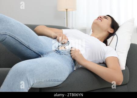 Young woman struggling to squeeze into tight jeans while lying on sofa at home Stock Photo