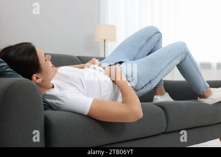 Young woman struggling to squeeze into tight jeans while lying on sofa at home Stock Photo