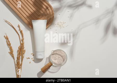 Wooden-handled glass bowl filled with rice bran powder. Unbranded facial cleanser tube, rice, and a domed podium on a white backdrop with soft shadows Stock Photo