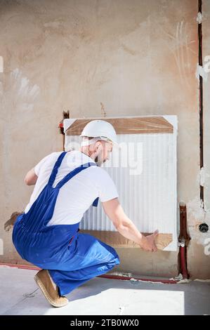 Back view of man central heating technician installing heating radiator on the wall. Man wearing safety construction helmet and work overalls while working on home renovation. Stock Photo