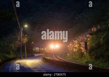A yellow diesel train passes through the village at night with its lights on. Along the Pingxi line, there are river valleys, potholes and waterfalls. Stock Photo