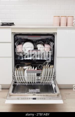 Open built-in dishwasher machine with clean cutlery, dishes, plates in white modern kitchen. front view Stock Photo
