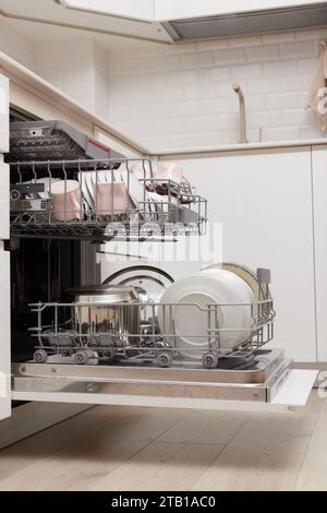 Open built-in dishwasher machine with clean cutlery, dishes, plates in white modern kitchen. side view Stock Photo