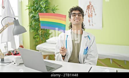 Young hispanic man doctor using laptop holding rainbow flag at clinic Stock Photo