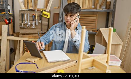 Experienced young hispanic carpenter multitasking, talking on a smartphone while using a touchpad in a busy carpentry workshop Stock Photo