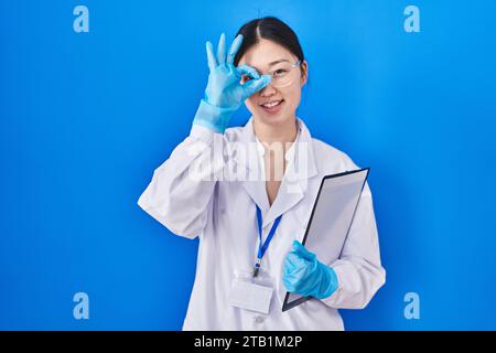 Chinese young woman working at scientist laboratory doing ok gesture with hand smiling, eye looking through fingers with happy face. Stock Photo