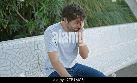 Serious young hispanic man, rocking a blond beard, combating stress sitting on city park bench. his casual, cool expression concealing worries under s Stock Photo