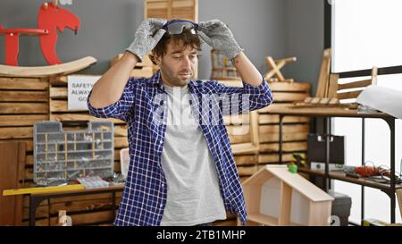 Handsome blond, bearded young man – a serious, professional carpenter wearing safety glasses, standing amid his timber and furniture in indoor carpent Stock Photo