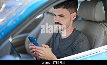 Young hispanic man using smartphone sitting on car at street Stock Photo