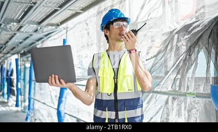 Young hispanic man architect talking on walkie-talkie using laptop at construction place Stock Photo