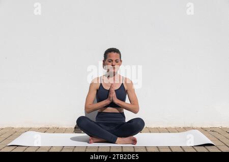 Woman sitting by a white wall on a yoga mat, meditating in the morning sun. Stock Photo