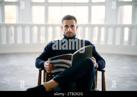 Young handsome man reading magazine seated comfortably in chair in modern white room. Relaxing concept Stock Photo