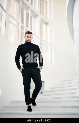 Fashionable man in a sleek black turtleneck descends a white modern staircase, exuding confidence and style. Stock Photo