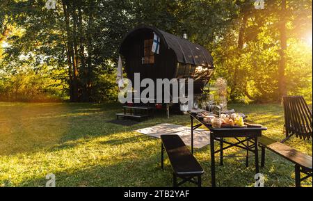 Finnish wooden sauna barrel with a table set with fresh food and wine glasses in a park at sunset Stock Photo