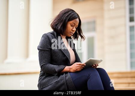 Young Black career woman sitting outside office with mobile device tablet Stock Photo