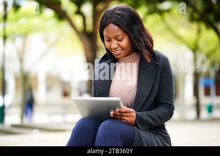 Young Black career woman smiling, sitting on bench in park with tablet Stock Photo