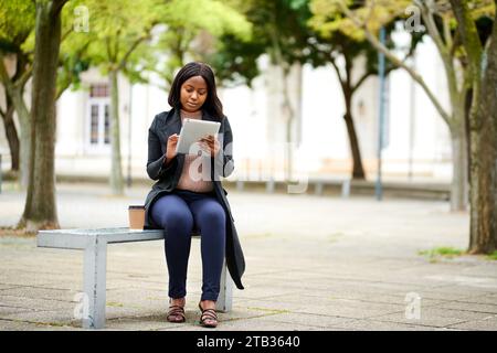 Young Black career woman sitting on outdoor bench with tablet, smiling Stock Photo
