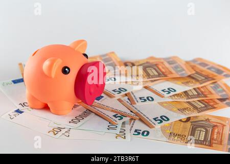 A pink pig piggy bank stands on a table covered with large quantity of euro banknotes on a light background Stock Photo