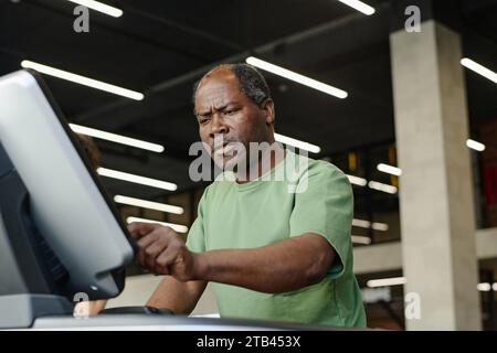 Medium low angle shot of elderly african american man pressing key on treadmill display at gym Stock Photo