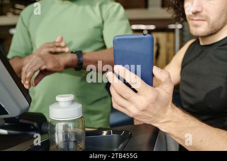 Unknown caucasian guy showing smartphone to unrecognizable black man standing on treadmill at gym Stock Photo