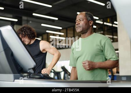 Elderly black man standing on treadmill at gym with caucasian gym goer working out on background Stock Photo