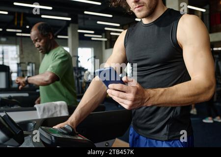 Unknown caucasian guy walking on treadmill with smartphone, black gym goer working out on background Stock Photo