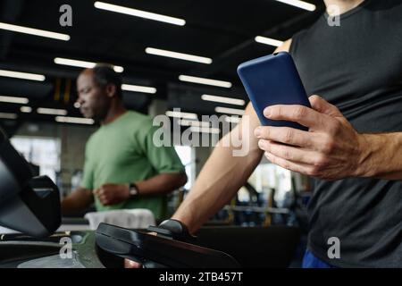 Unknown caucasian guy training at gym holding smartphone, elderly black man exercising on background Stock Photo