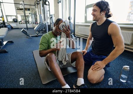 Senior black man sitting on floor at gym wiping forehead, caucasian instructor sitting next to him Stock Photo