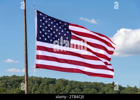 USA flag on flagpole flutters in the wind with red and white stripes and stars above a green forest and white clouded blue sky Stock Photo