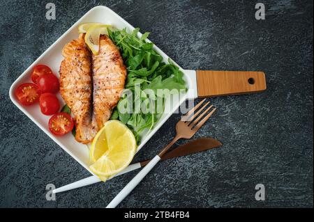 Grilled Butterfly Salmon Steak with cherry tomatoes and arugula salad on a white square plate. Close-up. Top view, flat lay, copy space. Stock Photo