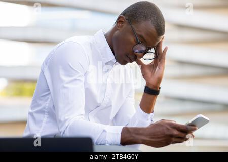 Focused black businessman in white shirt using smartphone while sitting outdoors Stock Photo