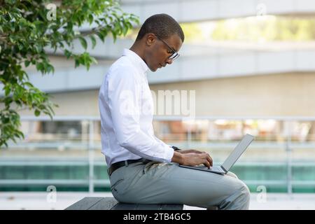 Concentrated black businessman in glasses working on his laptop while sitting outdoors Stock Photo
