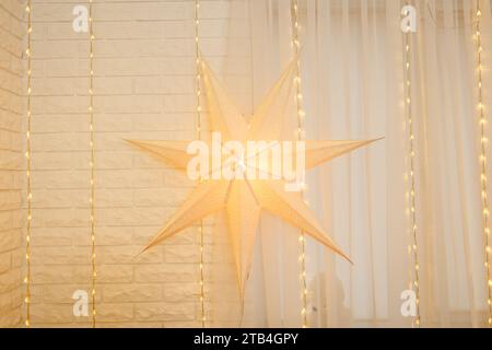 Christmas star on golden lights background on wall in living room. Large paper star glows against the backdrop of New Year garland. Atmospheric festiv Stock Photo