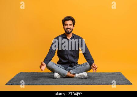 Relaxed young indian man in casual clothes meditating alone, sitting cross-legged on carpe Stock Photo