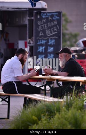 Two friends sharing companionship at a picnic table and beer garden while checking their mobile phones at sunset in downtown Milwaukee, Wisconsin, USA Stock Photo