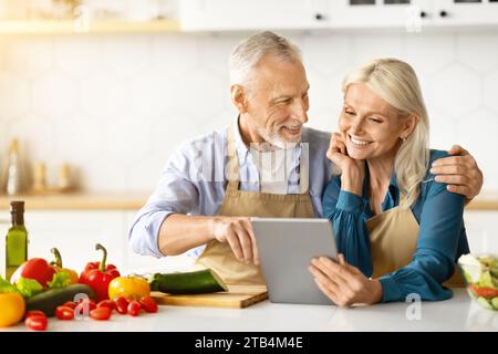 Cheerful Elderly Couple Using Digital Tablet While Cooking Healthy Lunch In Kitchen Stock Photo