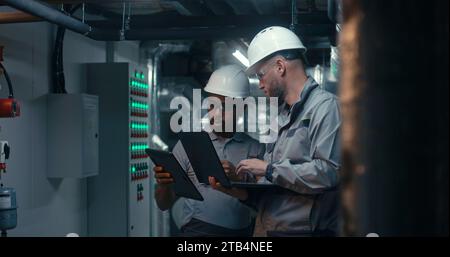 Young diverse team of factory specialists work on laptop and tablet computer. Professional engineers discuss industrial manufacturing technology on modern plant. Teamwork in heavy industry concept. Stock Photo